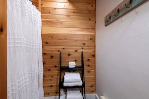 Un bagno con pareti rivestite in legno, una mensola in metallo nero con asciugamani piegati e una tenda bianca.