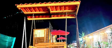 Un columpio de madera con techo, rodeado de rocas y un edificio con luces al fondo.