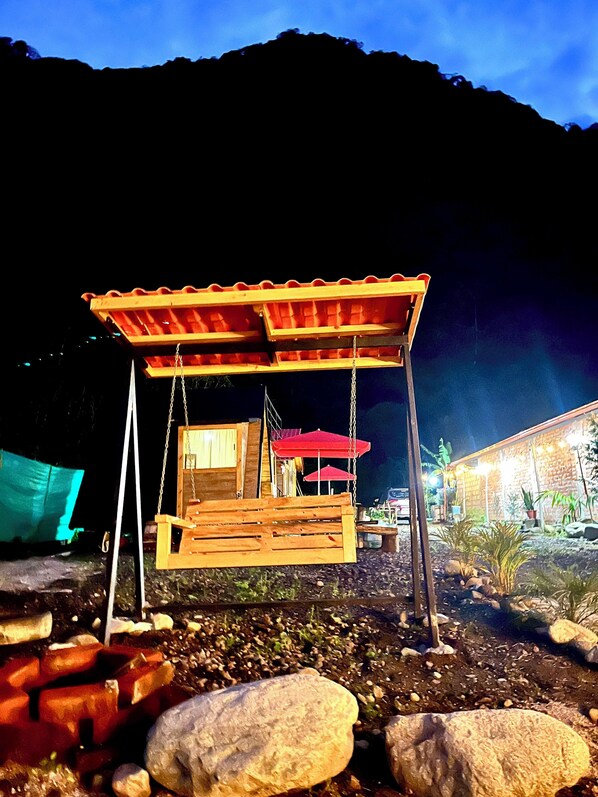A wooden swing set with a roof, surrounded by rocks and a building with lights in the background.
