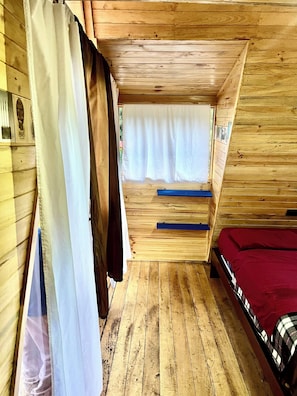 A wooden interior with a bed, a window with curtains, and a wooden floor. - "Here, you can forget about the world." A little house, mountains, a river, fishing, birds, and above all, art (Río Verde)
