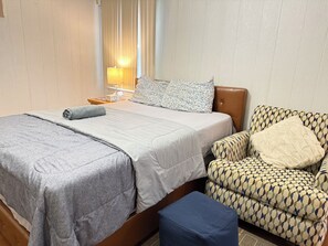 Ein ordentlich bezogenes Bett mit grauer Decke und Kissen, ein gemustertes Sessel und ein Nachttisch mit Lampe.