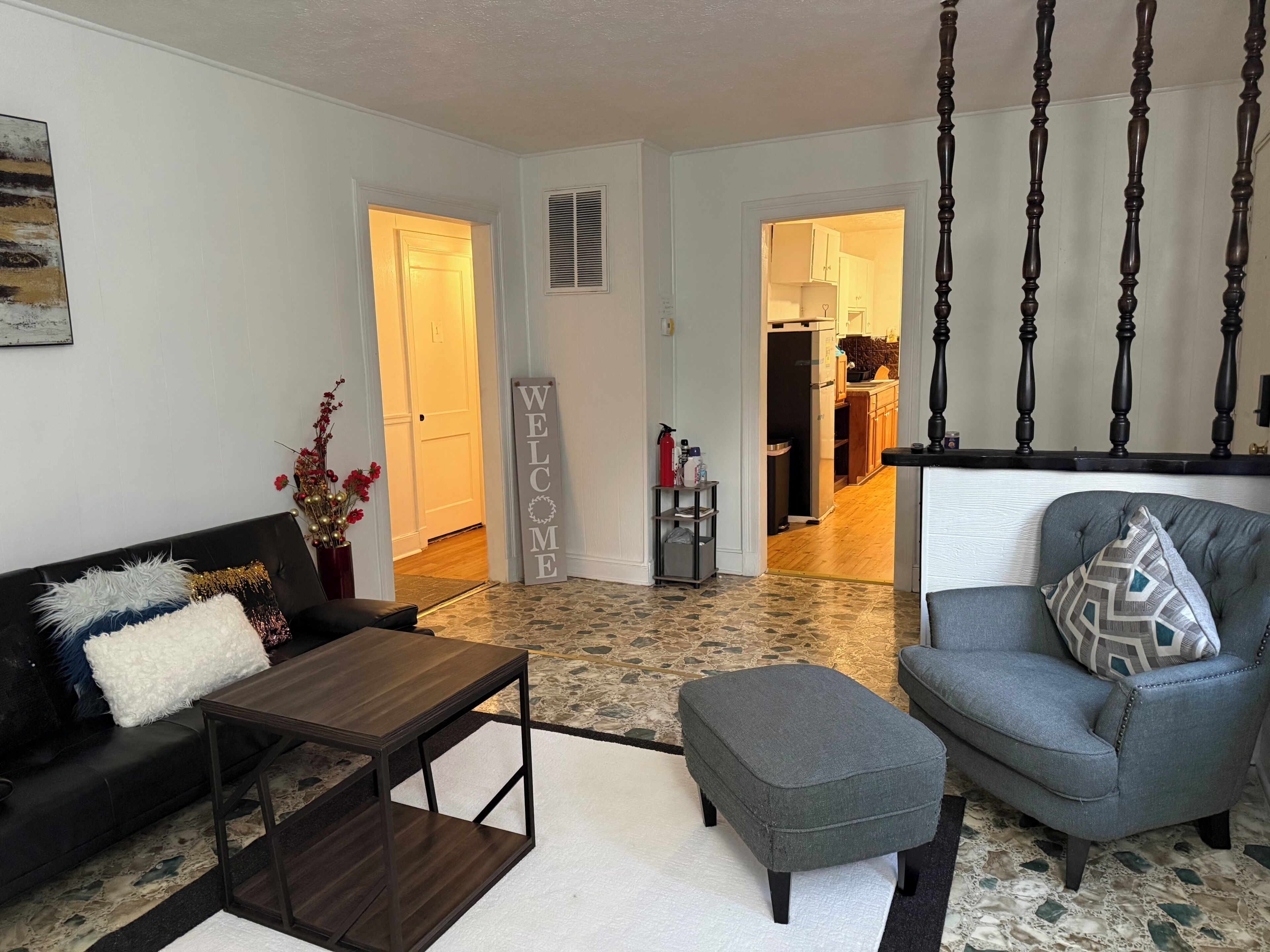 A living room with a sofa, ottoman, coffee table, and a chair with a cushion. A welcome sign is visible in the hallway.