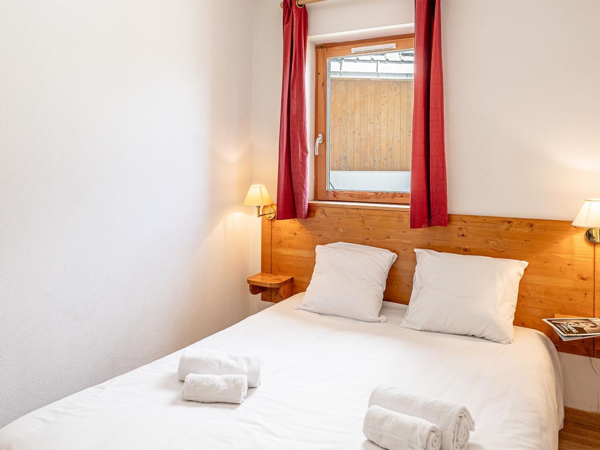 A hotel room with a wooden headboard, a window with red curtains, two bedside lamps, and a neatly made bed with white linens and towels.