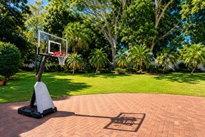 Un terrain de basket avec un panier, entouré d’un chemin pavé de briques et d’une végétation luxuriante.