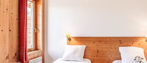 Una habitación con dos camas, cabeceros de madera, un ventanal con cortina roja y una pequeña mesa de madera con lámpara.