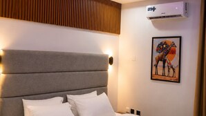 A modern hotel room with a large bed, a headboard with built-in lighting, an air conditioning unit, and a wall-mounted artwork.