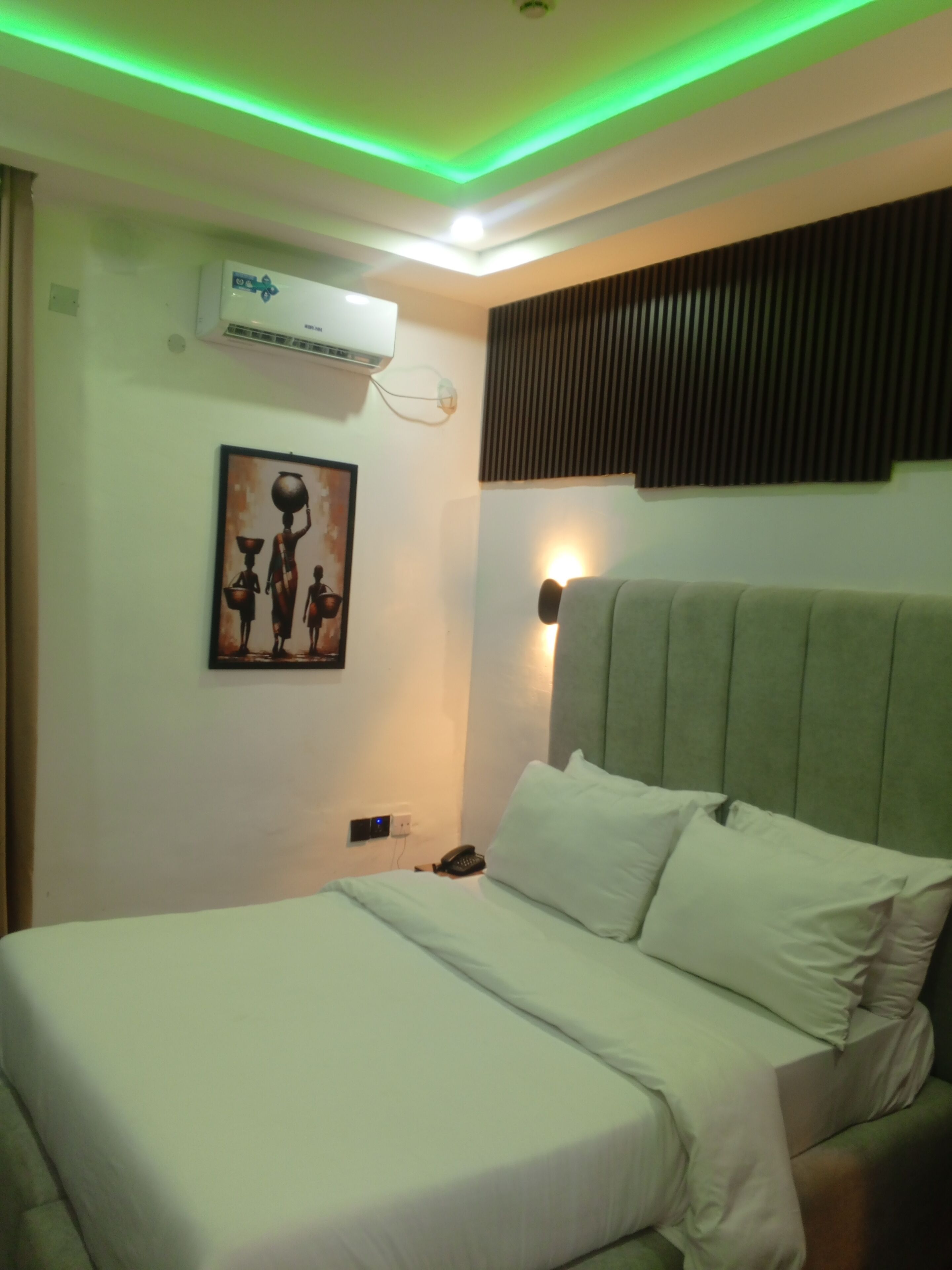 A hotel room with a bed, a wall-mounted air conditioner, a painting, a bedside lamp, and a telephone.