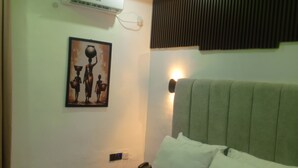 A hotel room with a bed, a wall-mounted air conditioner, a painting, a bedside lamp, and a telephone.