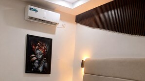 A hotel room with a bed, white bedding, an air conditioning unit, a wall-mounted light, and a framed artwork.