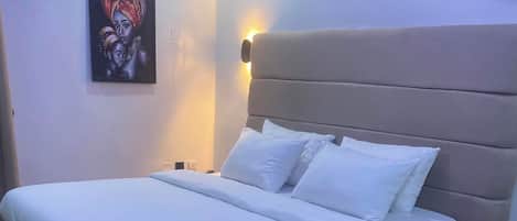 Una cama con cabecera, ropa de cama blanca y una lámpara fijada a la pared.