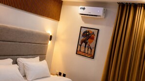 A hotel room with a bed, an air conditioning unit, a wall-mounted artwork, and a window with curtains.