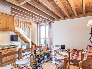 Une cuisine chaleureuse avec des poutres en bois, un coin repas avec une table dressée pour deux personnes, et un escalier menant à l’étage supérieur.