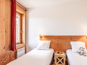 Ein Zimmer mit zwei Betten, Holz-Kopfteilen, einem Fenster mit rotem Vorhang und einem kleinen Holztisch mit Lampe.