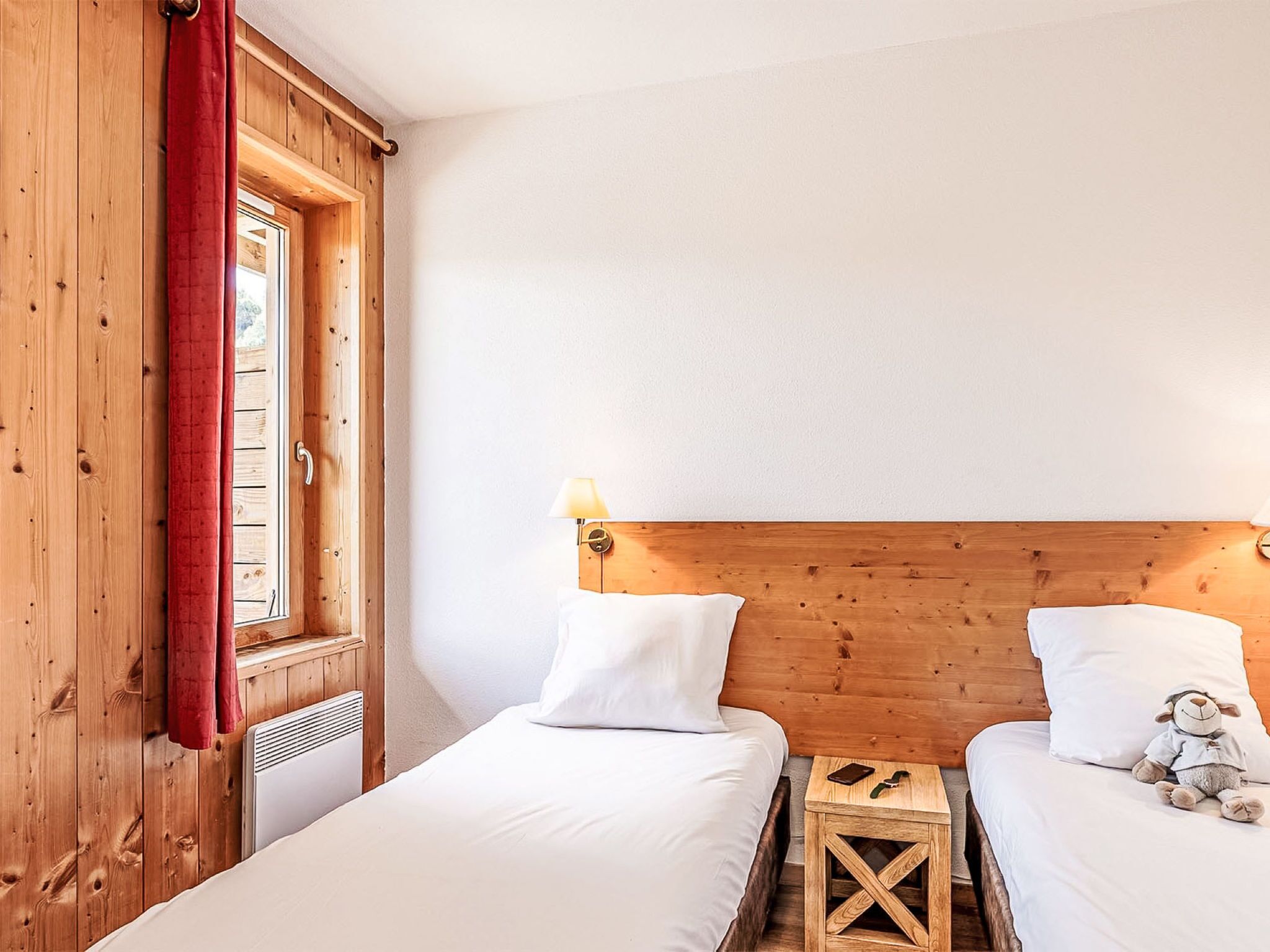 Una habitación con dos camas, cabeceros de madera, un ventanal con cortina roja y una pequeña mesa de madera con lámpara.