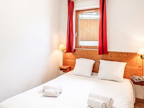 Ein Hotelzimmer mit einem Holzkopfteil, einem Fenster mit roten Vorhängen, zwei Nachttischen mit Lampen und einem ordentlich bezogenen Bett mit weißen Leinen und Handtüchern.
