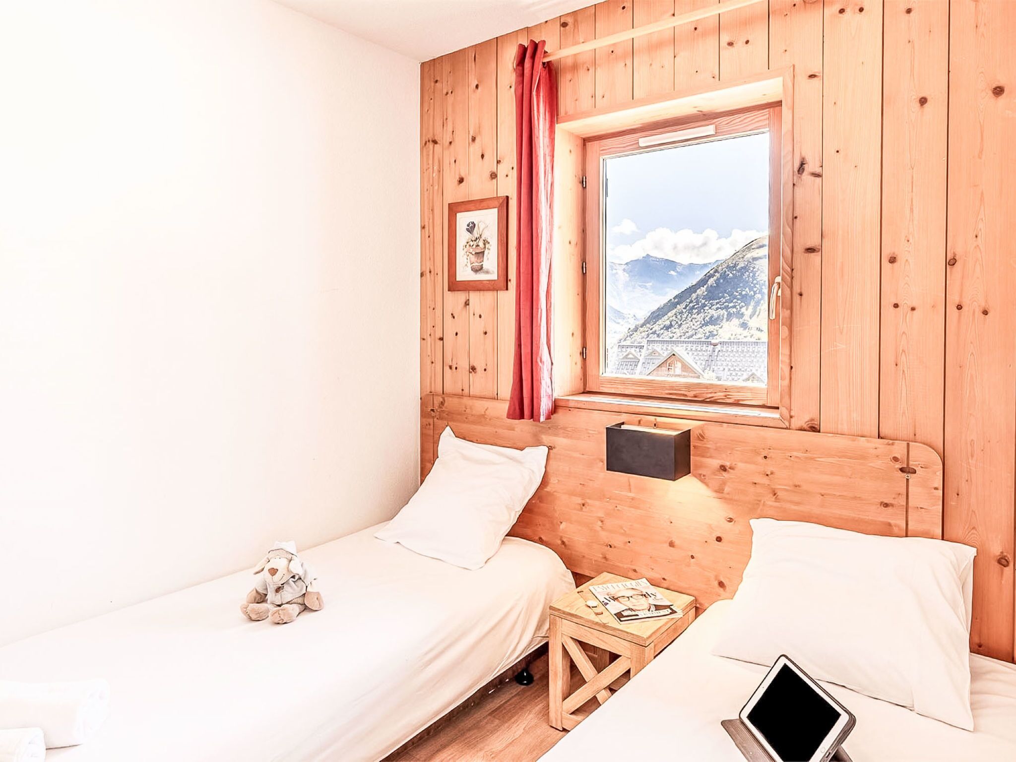 A wooden room with two beds, a window with a view of mountains, a small table with a lamp, and a stuffed toy on one bed.
