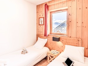 Une pièce en bois avec deux lits, une fenêtre donnant sur les montagnes, une petite table avec une lampe et un jouet en peluche sur l’un des lits.