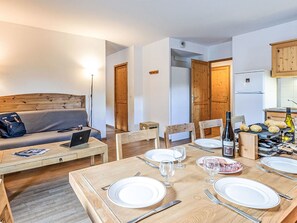 Een eetgedeelte met een houten tafel gedekt voor vier personen, een laptop op een bank en een kitchenette met een koelkast en fornuis.