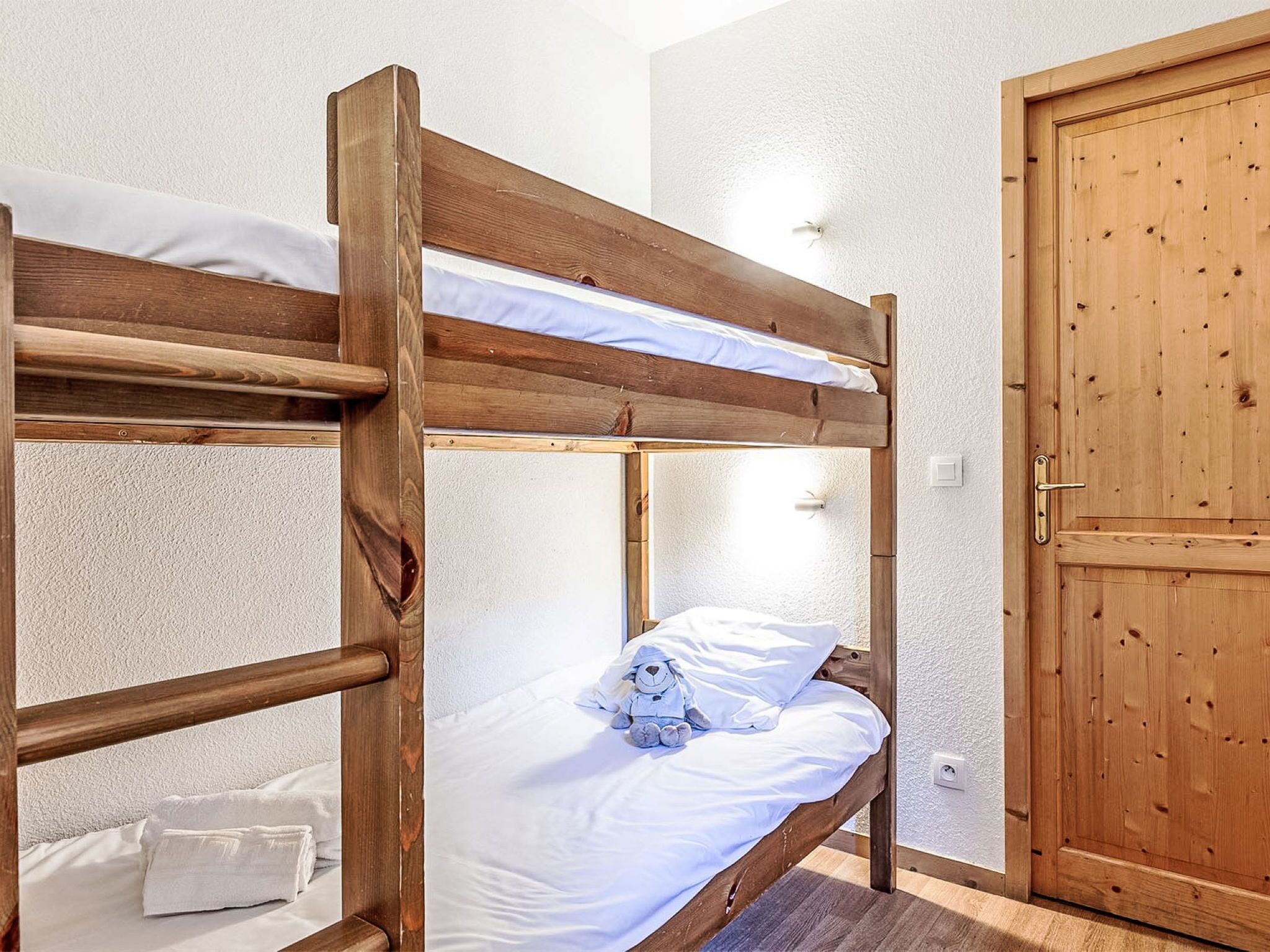 Una cama litera con estructura de madera, ropa de cama blanca y un osito de peluche en la cama inferior.