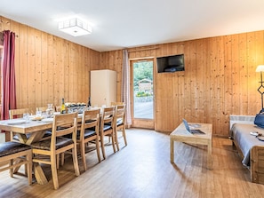 A room with wooden walls, a dining table with chairs, a sofa, a TV mounted on the wall, and a window with a view of greenery. - Les Fermes de Saint Sorlin by Interhome (Saint Sorlin d'Arves)