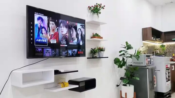 Ein modernes Wohnzimmer mit einem an der Wand befestigten Flachbildfernseher, der eine Auswahl an Filmen zeigt. Weiße Regale mit Dekoration und einem Topf mit Pflanze sind ebenfalls zu sehen.
