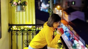 Eine Person mit gelber Kapuzenjacke sitzt auf einem Balkon mit Geländer und hält eine rote Tasse.