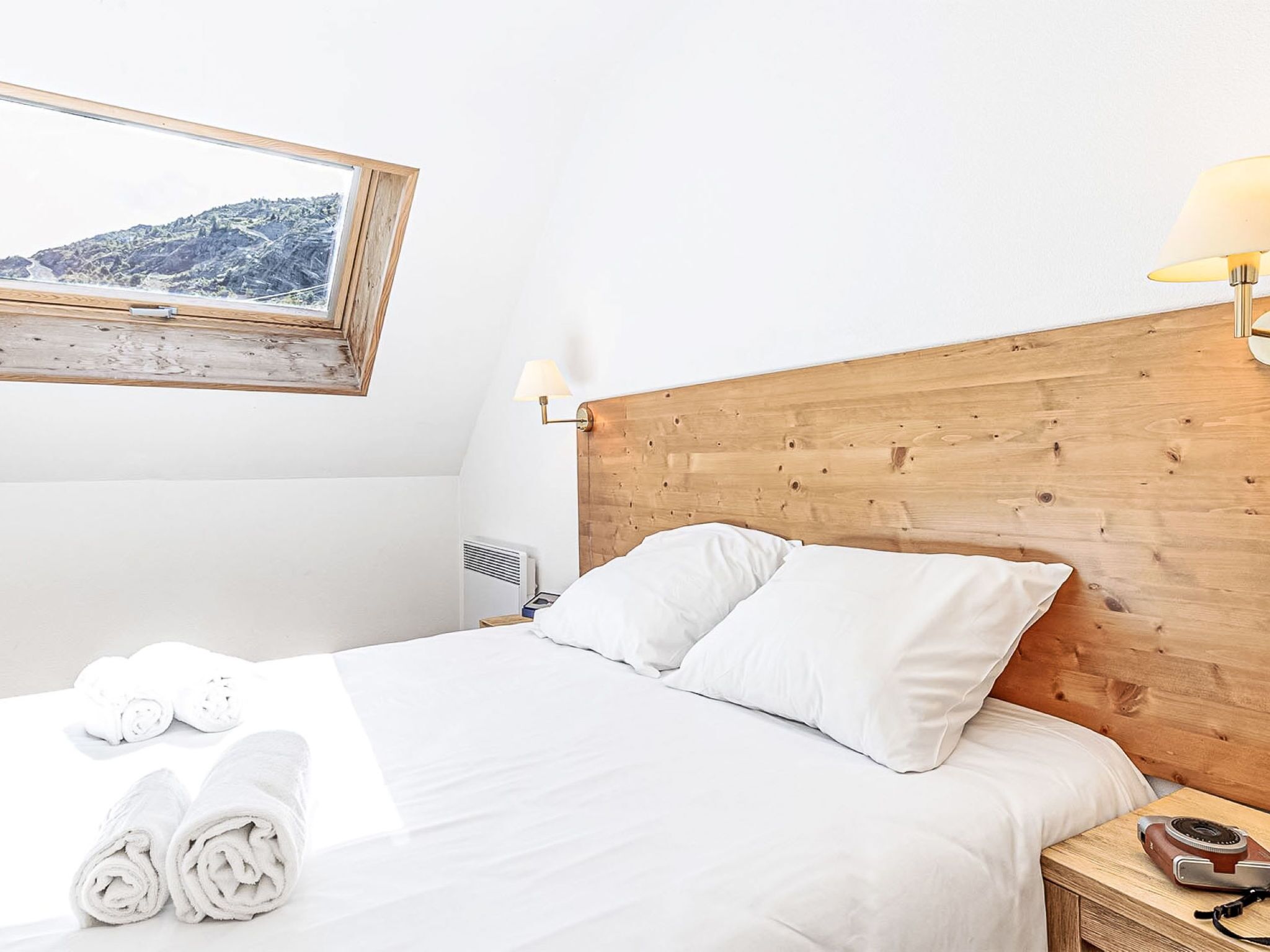 A hotel room with a wooden headboard, white bedding, a rolled towel, a bedside lamp, and a view of a mountainous landscape through a window.