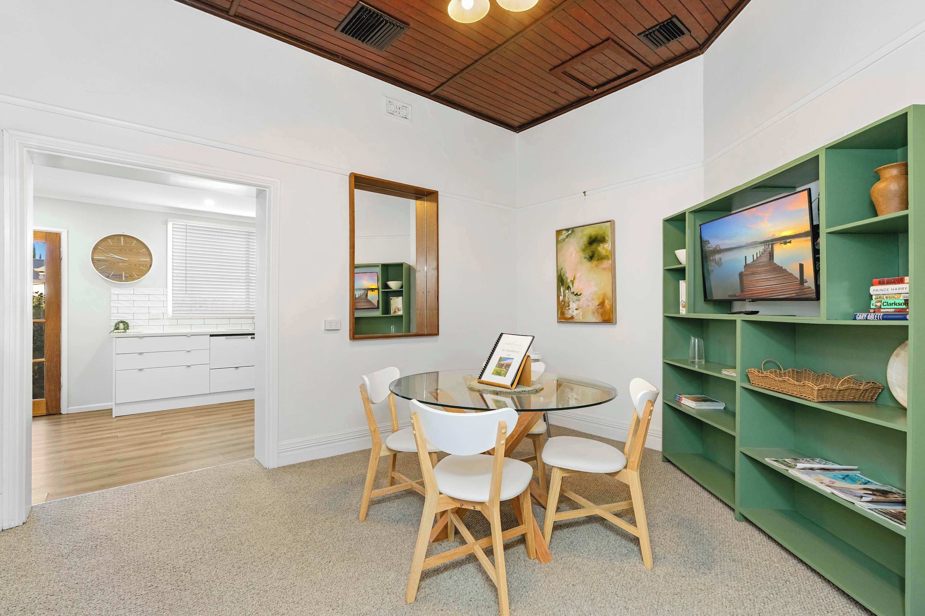 A modern interior with a round glass table surrounded by white chairs, a green bookshelf, and a flat-screen TV.