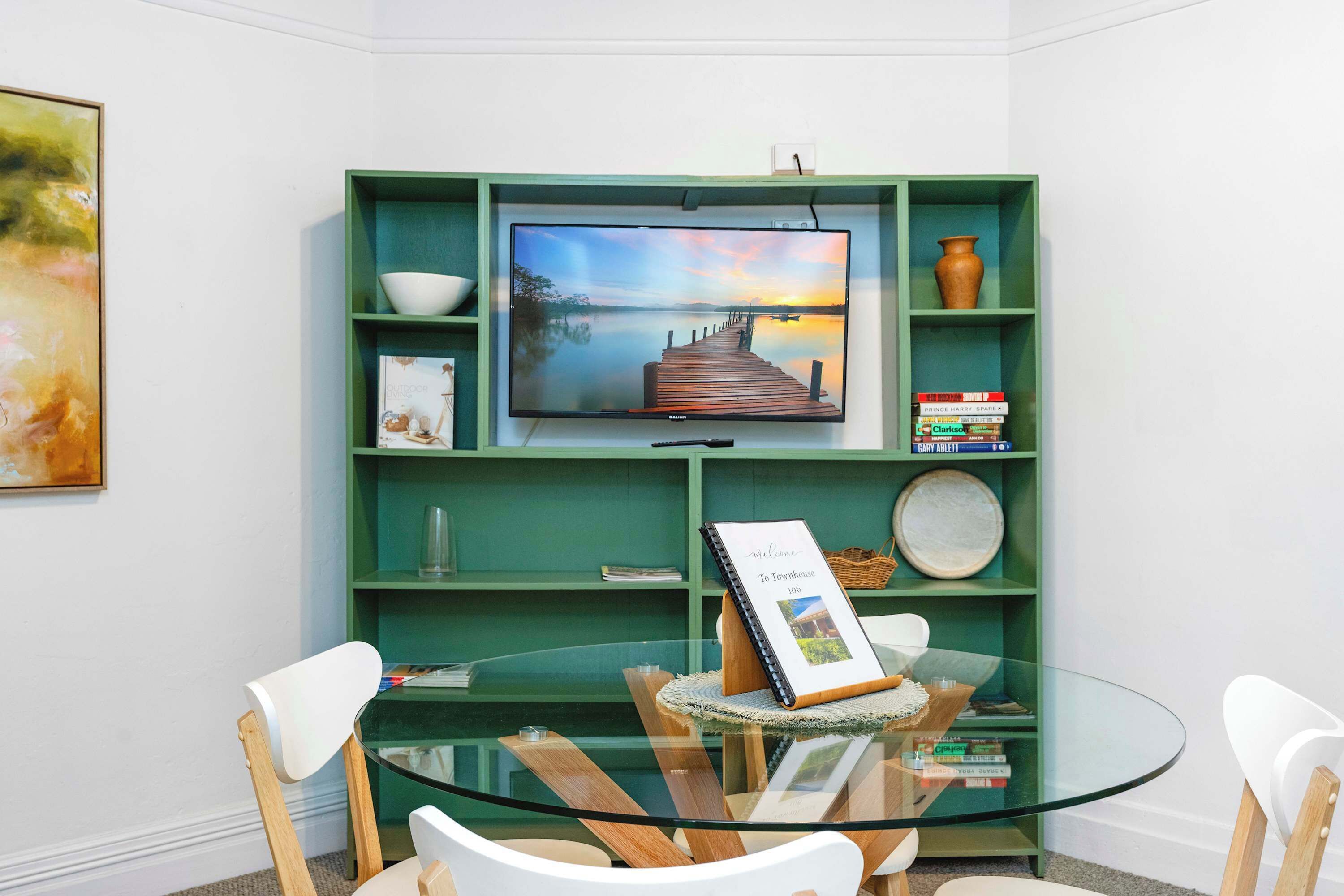 Ein modernes Wohnzimmer mit Glastisch, weißen Stühlen, einer grünen Unterhaltungseinheit und einem Flachbildfernseher, sowie einem gerahmten Willkommen-Schild.