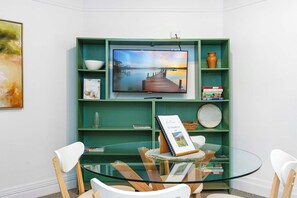 Un salon moderne avec une table en verre, des chaises blanches, un meuble de rangement vert abritant un téléviseur à écran plat et un panneau d’accueil encadré.