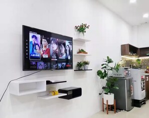 A modern living room with a flat-screen TV mounted on the wall displaying a selection of movies. There are white shelves with decorative items and a potted plant. - Localview Balcony-Trainstreet -Netflix-King Bed-Guide book (Hà Nội)