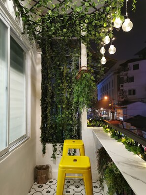 A balcony with a white railing, a yellow stool, and hanging plants. - Localview Balcony-Trainstreet -Netflix-King Bed-Guide book (Hà Nội)