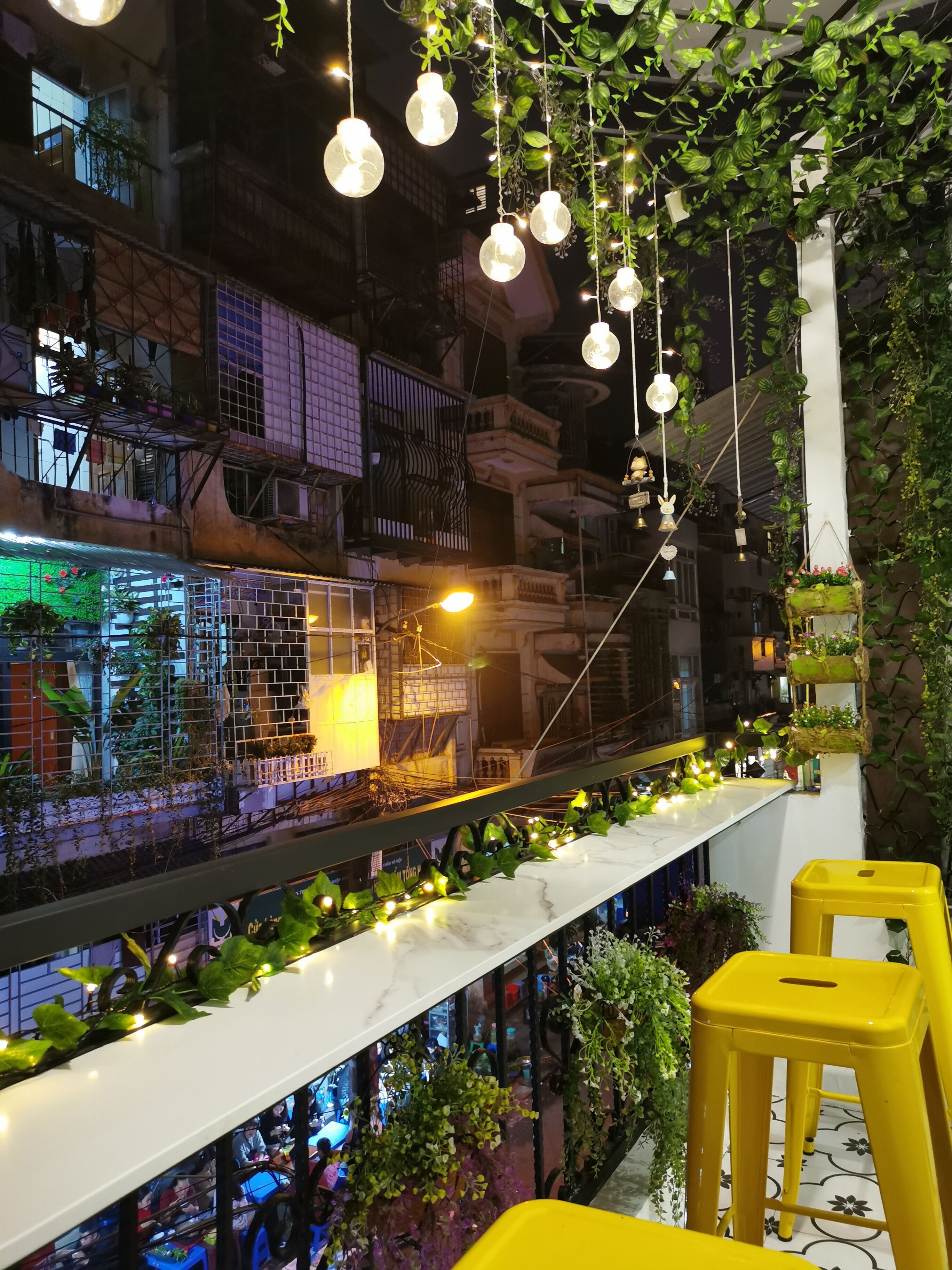 A balcony with hanging lights, greenery, and yellow stools.