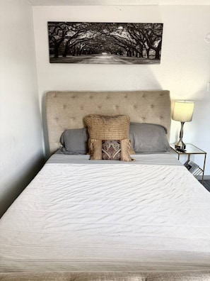 Een bed met hoofdbord, twee kussens, een decoratief kussen en een aan de muur hangend schilderij.