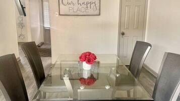 A dining area with a glass-top table surrounded by four chairs, a vase with red flowers, and a sign on the wall that reads 'Our happy PLACE'.