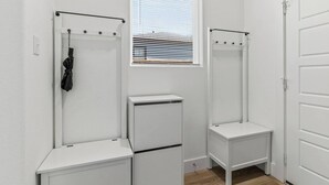 Une petite pièce blanche, avec une seule fenêtre, une porte et un porte-manteau sur lequel est accroché un vêtement noir.