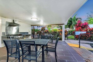 Ein Essbereich im Freien mit Metallstühlen und -tischen, einer Grillstation und Blick auf einen angelegten Garten mit tropischen Pflanzen.