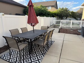 En udendørs terrasse med et bord, stole, en stribet måtte og et hvidt hegn.