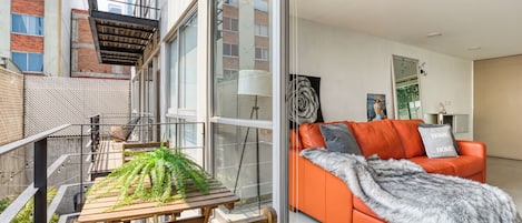 A modern living room with a glass door leading to a balcony, a wooden bench, and an orange sofa with a fur throw.