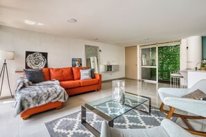 Een moderne woonkamer met een glazen salontafel, een oranje bank en uitzicht op een groen buitengebied.