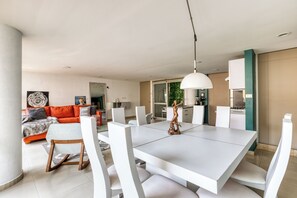 Een moderne eethoek met een witte tafel en stoelen, een hanglamp en zicht op de woonkamer met een bank en kussens.