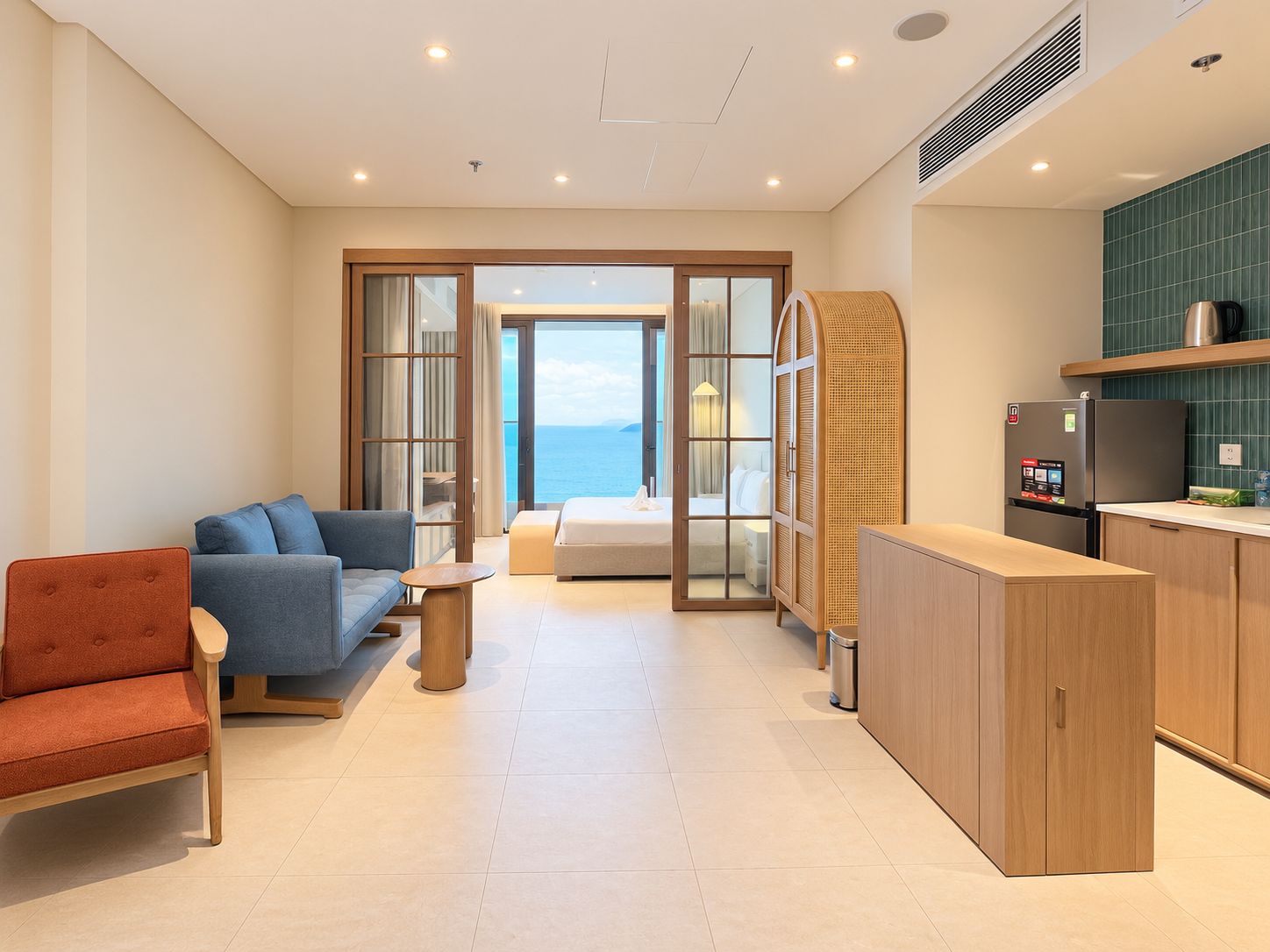 A modern hotel room with a sofa, a bed, a small table, a kitchen area, and a view of the sea through the sliding glass door.
