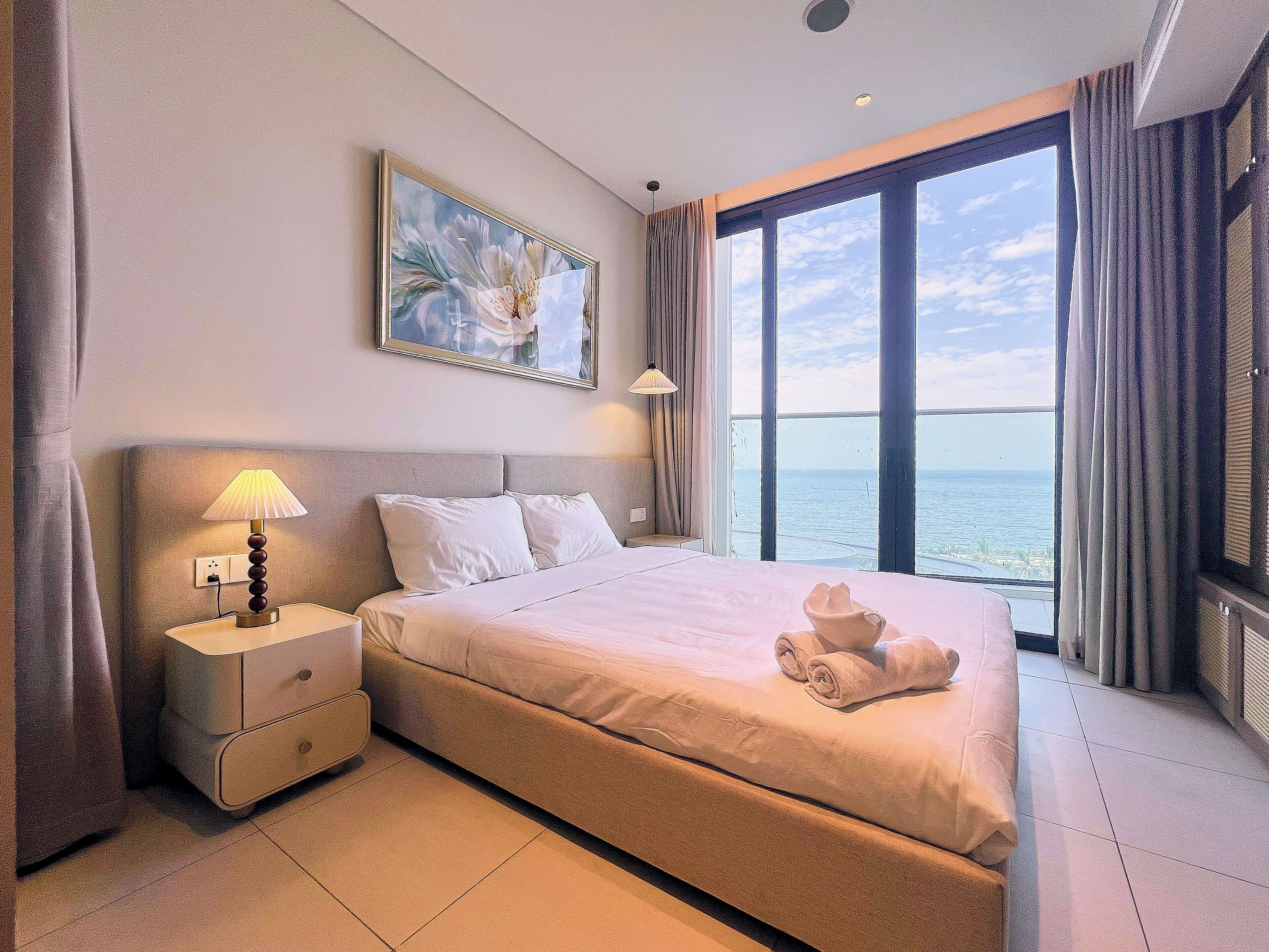 Una habitación de hotel con cama, mesita de noche, lámpara, un cuadro y vistas al mar.