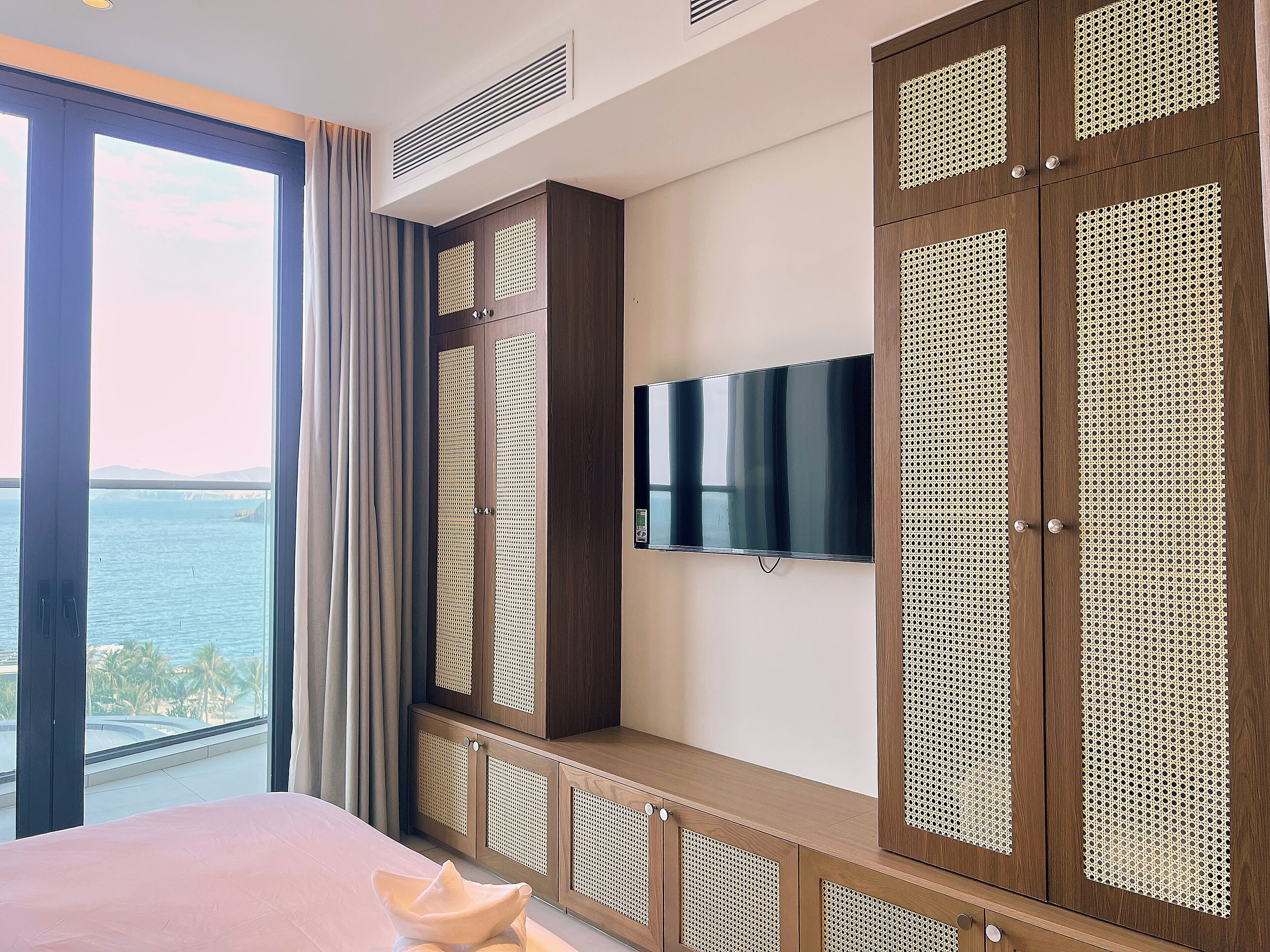 Un dormitorio con un ventanal grande, una cama, un televisor y un armario de madera con diseño tejido.