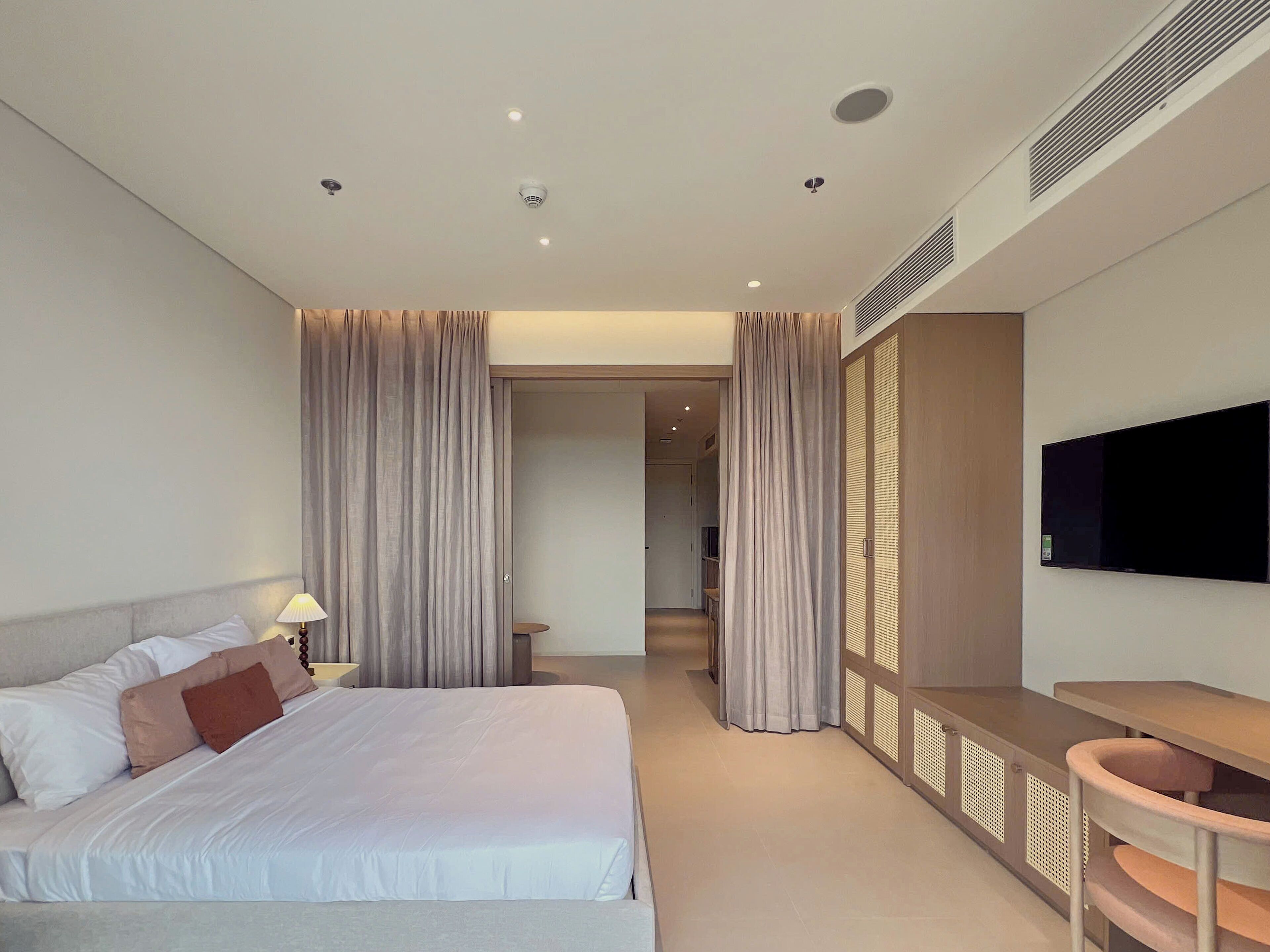 Una habitación de hotel moderna con cama, mesita de noche, lámpara, cortinas y televisor.