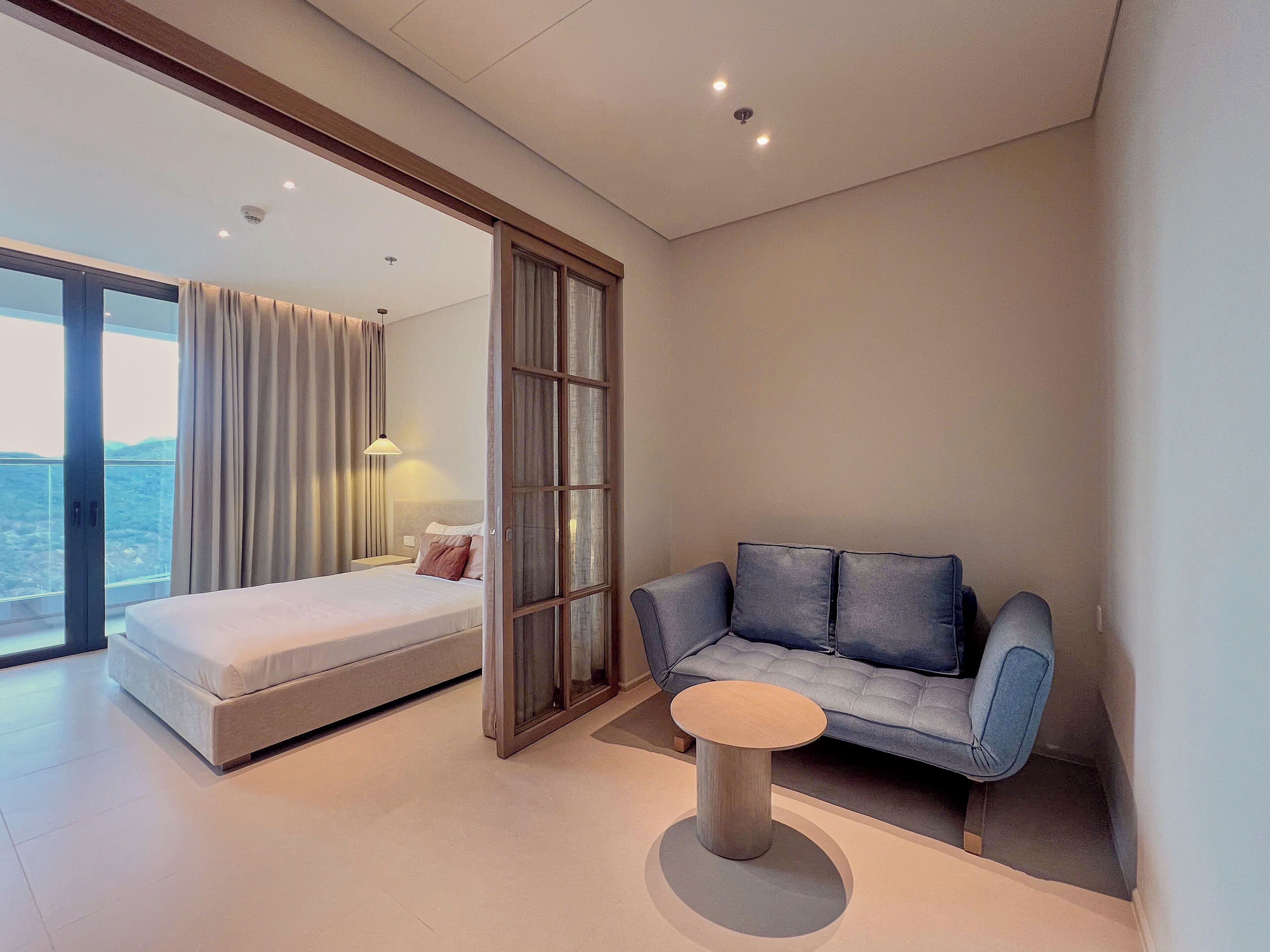 A modern hotel room with a bed, a sofa, a small table, and a large window offering a view.