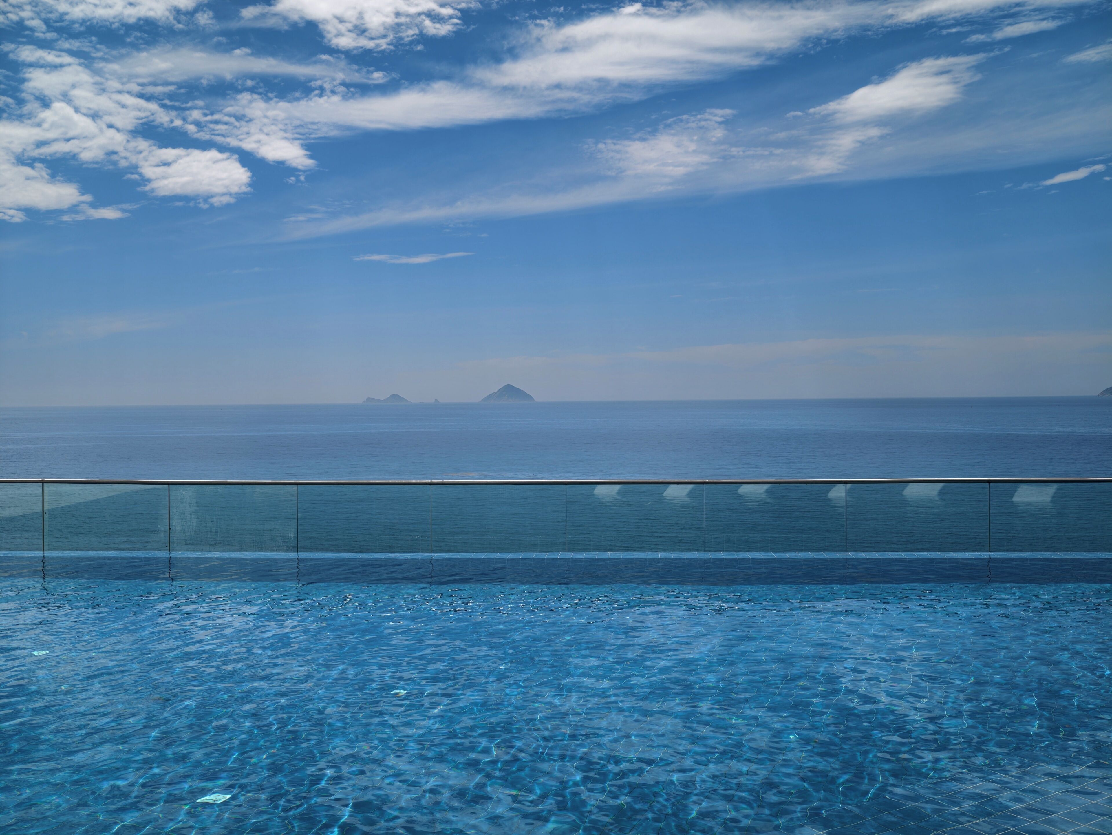 A clear blue sky with clouds, a calm sea, and a swimming pool with a glass barrier.