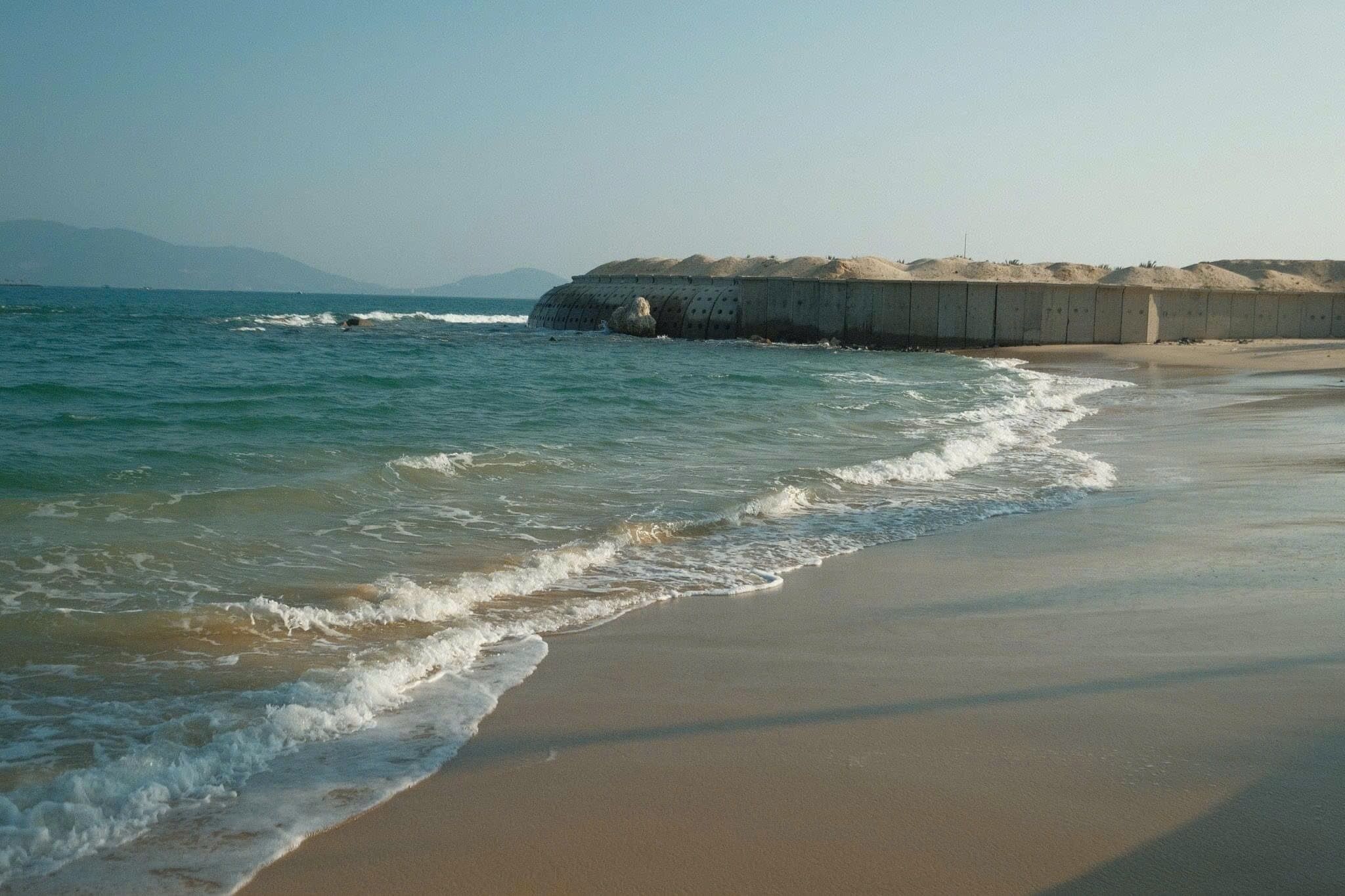 A beach with waves, a concrete barrier, and a clear sky.