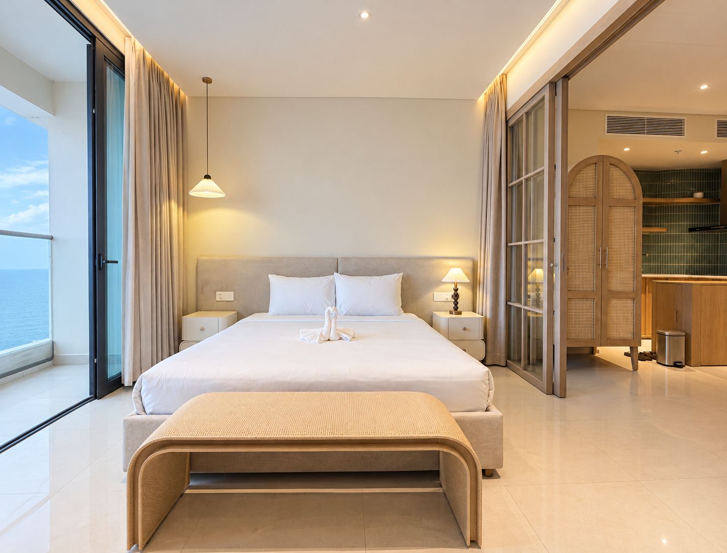 Quarto de hotel moderno com uma cama grande, um banco, uma mesa de cabeceira, uma luminária e vista para o mar.