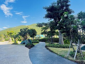 A paved pathway leading through a landscaped area with various trees and shrubs, a clear blue sky, and a hilly backdrop.